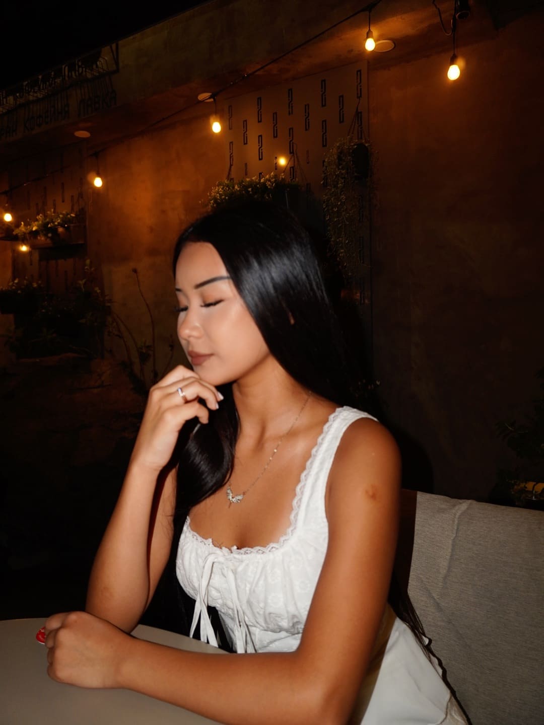With eyes closed and hand near her chin, a woman sits at a table in a white lace top, framed by hanging string lights.