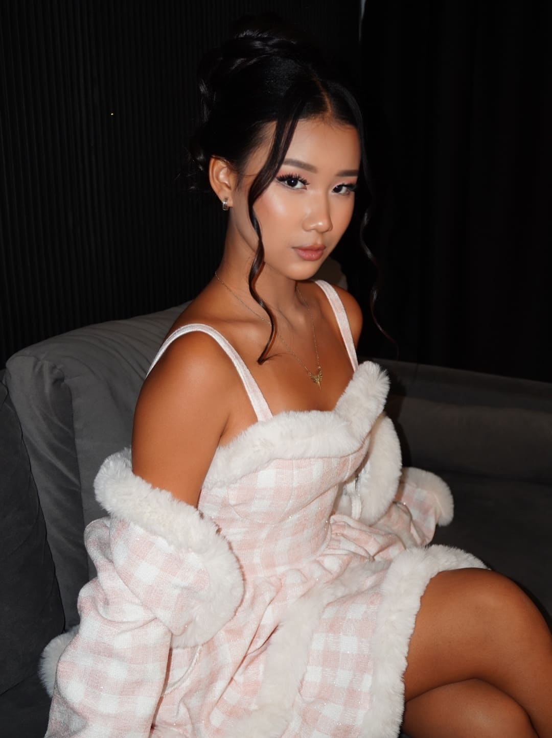 Assel sits on a gray sofa wearing a pink and white checkered dress with fur trim and a matching coat draped off her shoulders.