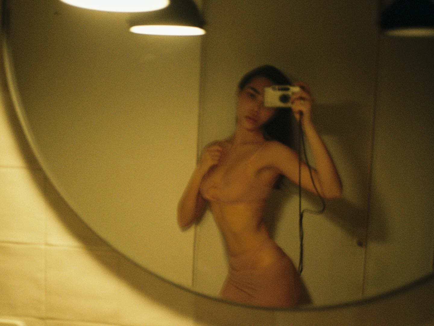 Mirror selfie in a dimly lit room, wearing a sheer nude slip dress that reveals the silhouette under warm light.