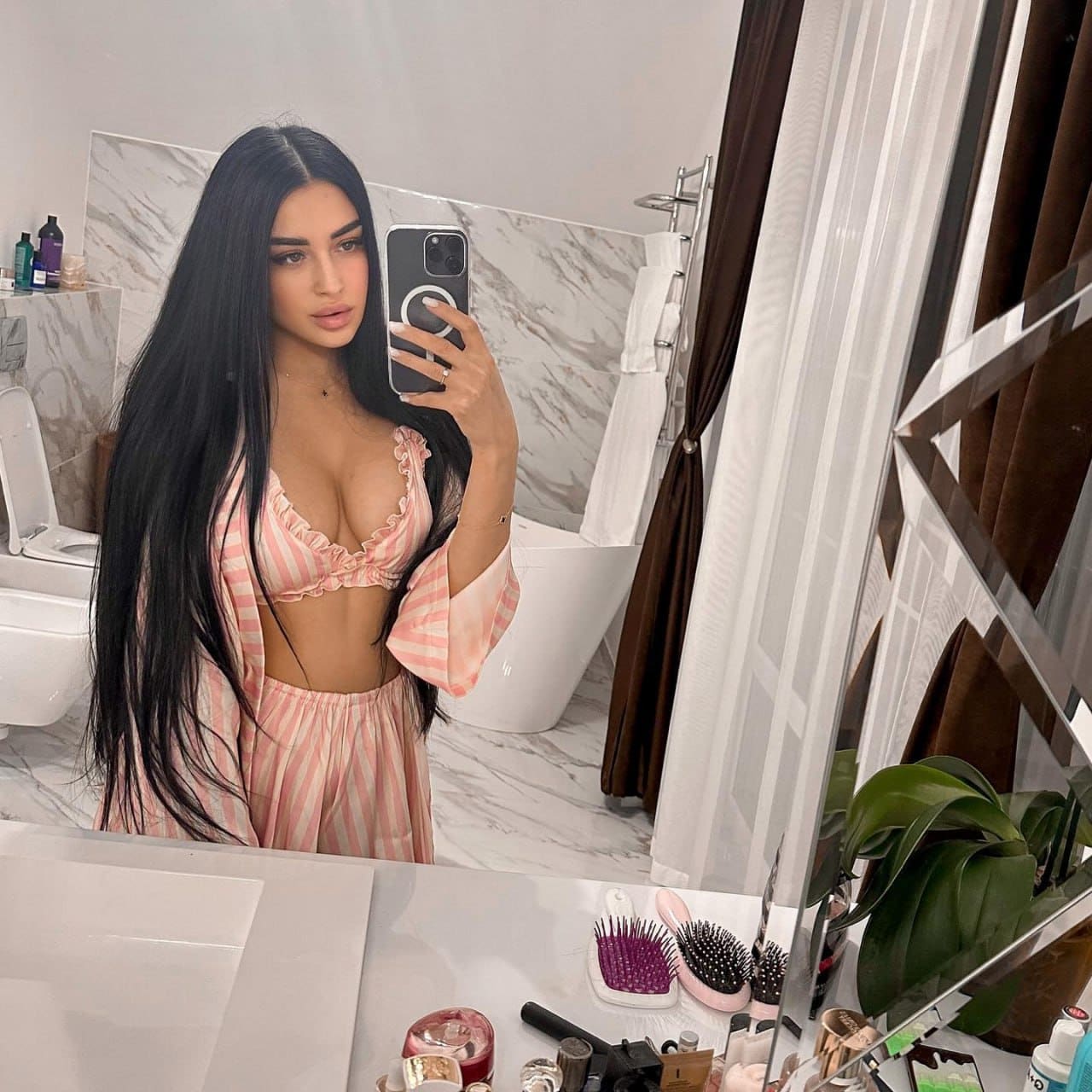 A woman takes a bathroom mirror selfie wearing a pink and white striped ruffled bra top and matching bottoms, showing an open midriff.