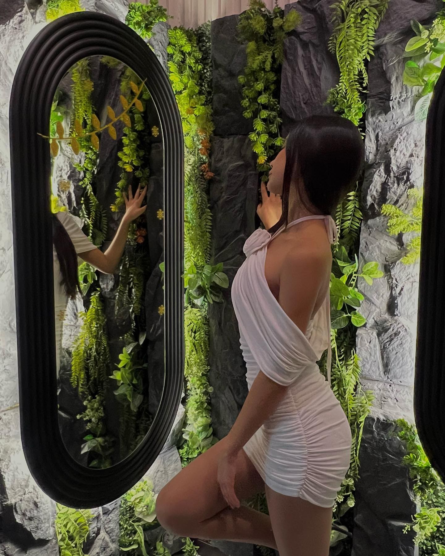 A woman stands in profile against a greenery wall and oval mirror, wearing a short white ruched halter dress with one leg raised.