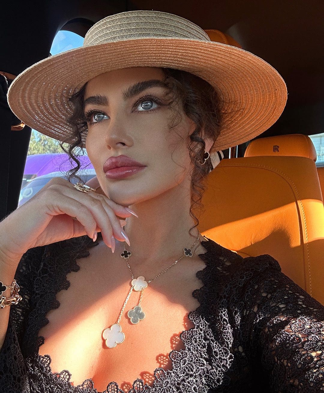 A woman in a straw hat and black lace top sits in a car with tan seats, hand near chin, wearing a clover necklace.