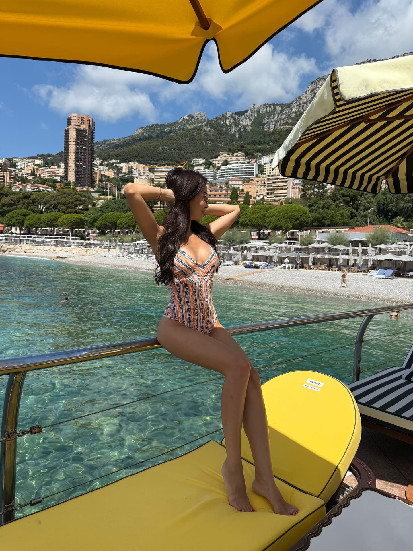 A woman sits on a yellow lounge chair in a patterned one-piece swimsuit, hands behind her head, with coastal mountains in view.