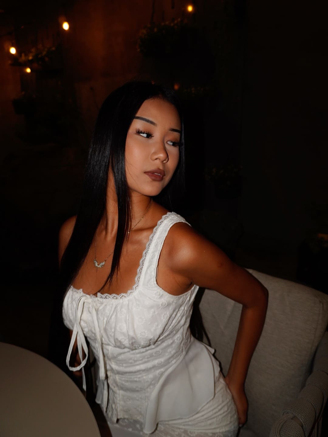 Seated in dim light, a woman with straight black hair wears a white lace-trimmed top with a front tie, looking off to the side.