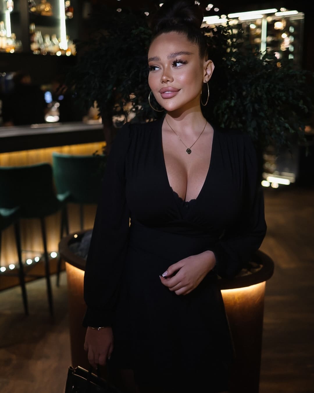 Woman with a high bun in a dim lounge, wearing a black long-sleeved dress with a deep V-neck, hoop earrings, and a pendant necklace.