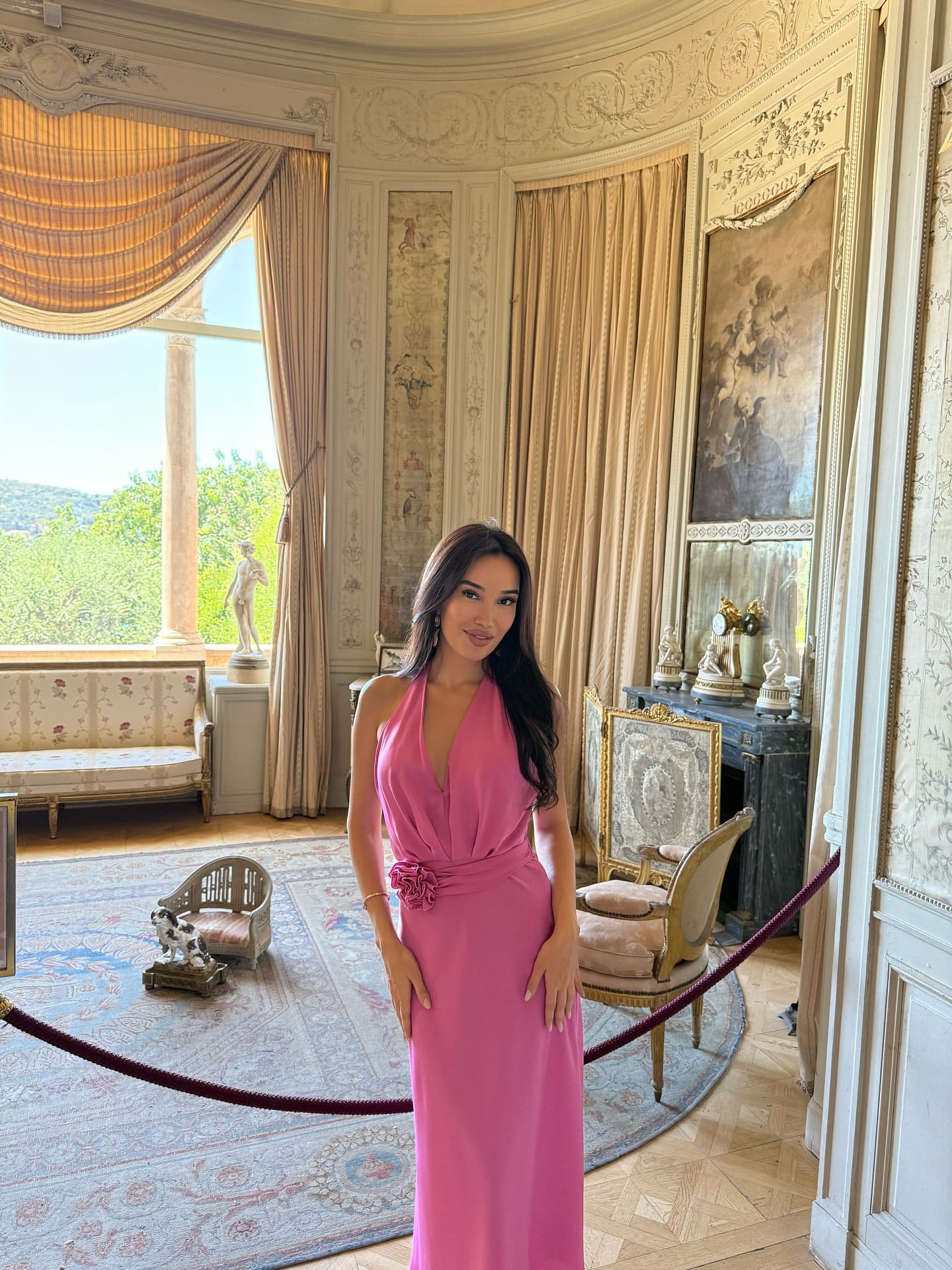 A woman stands in an ornate room wearing a floor-length pink halter gown with a plunging neckline and floral waist detail.