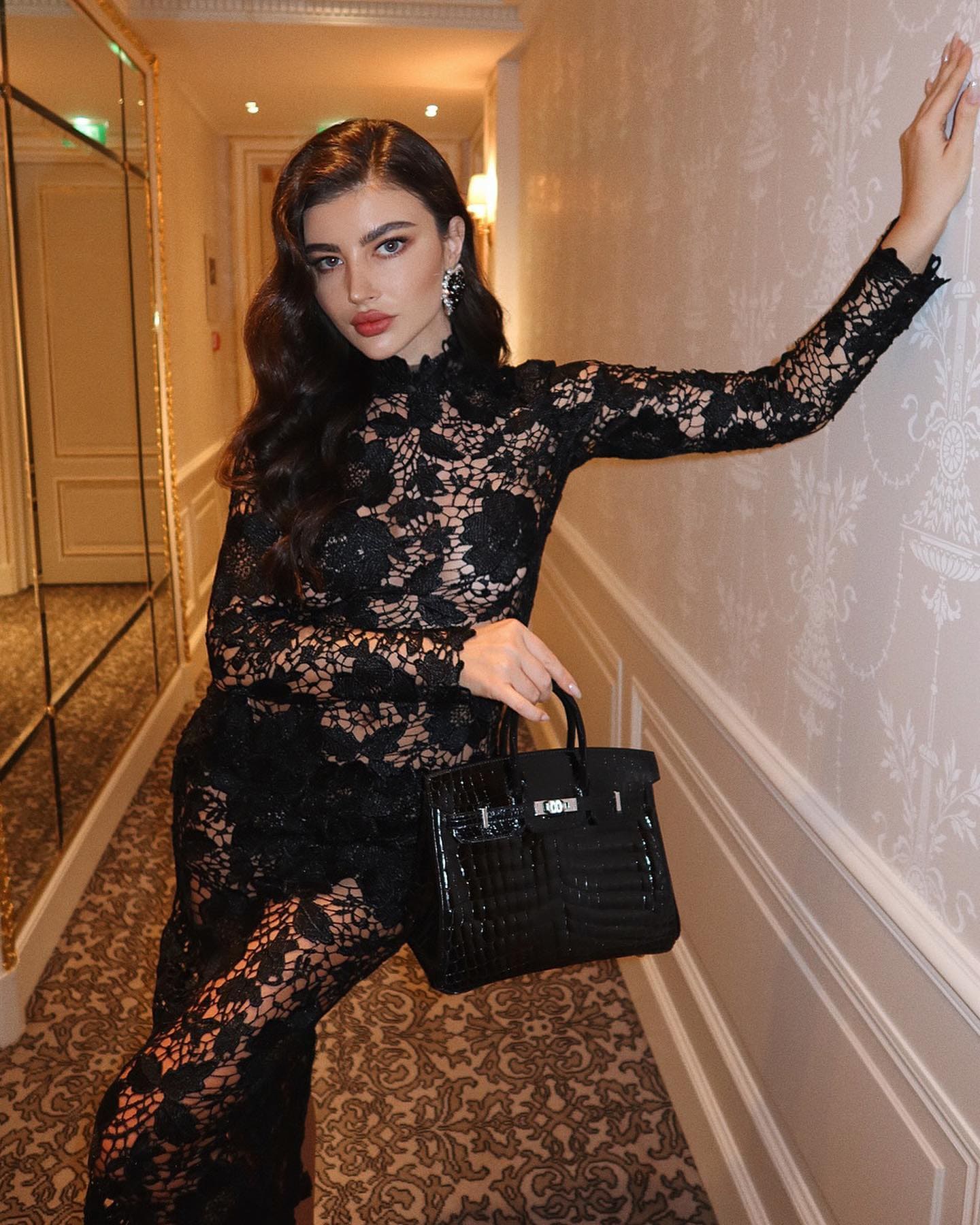 A woman stands in a hallway in a sheer floor-length black lace dress, one arm raised against the wall, holding a black bag.