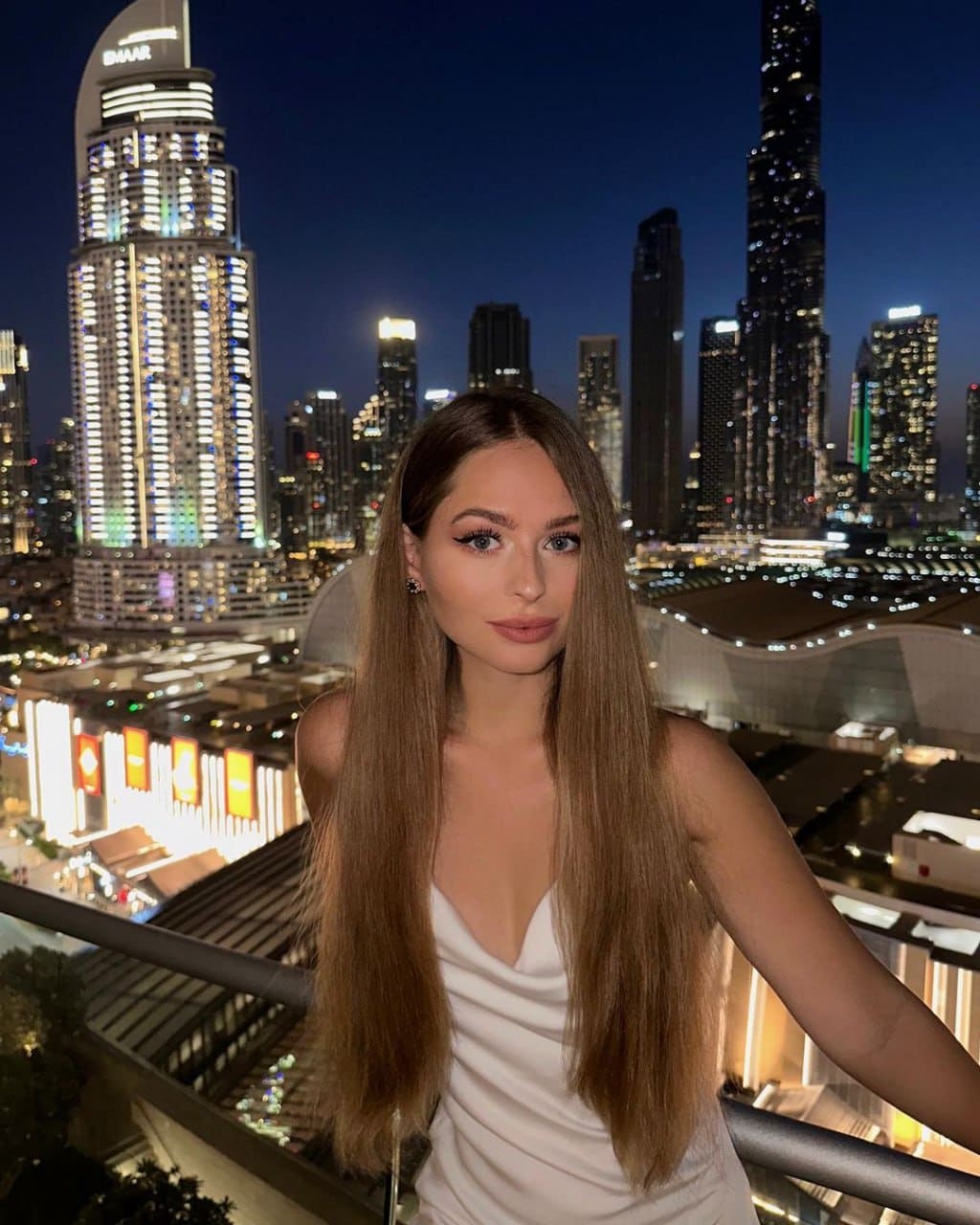 Woman in a white deep-V dress stands on a balcony at night, with an illuminated city skyline behind her.