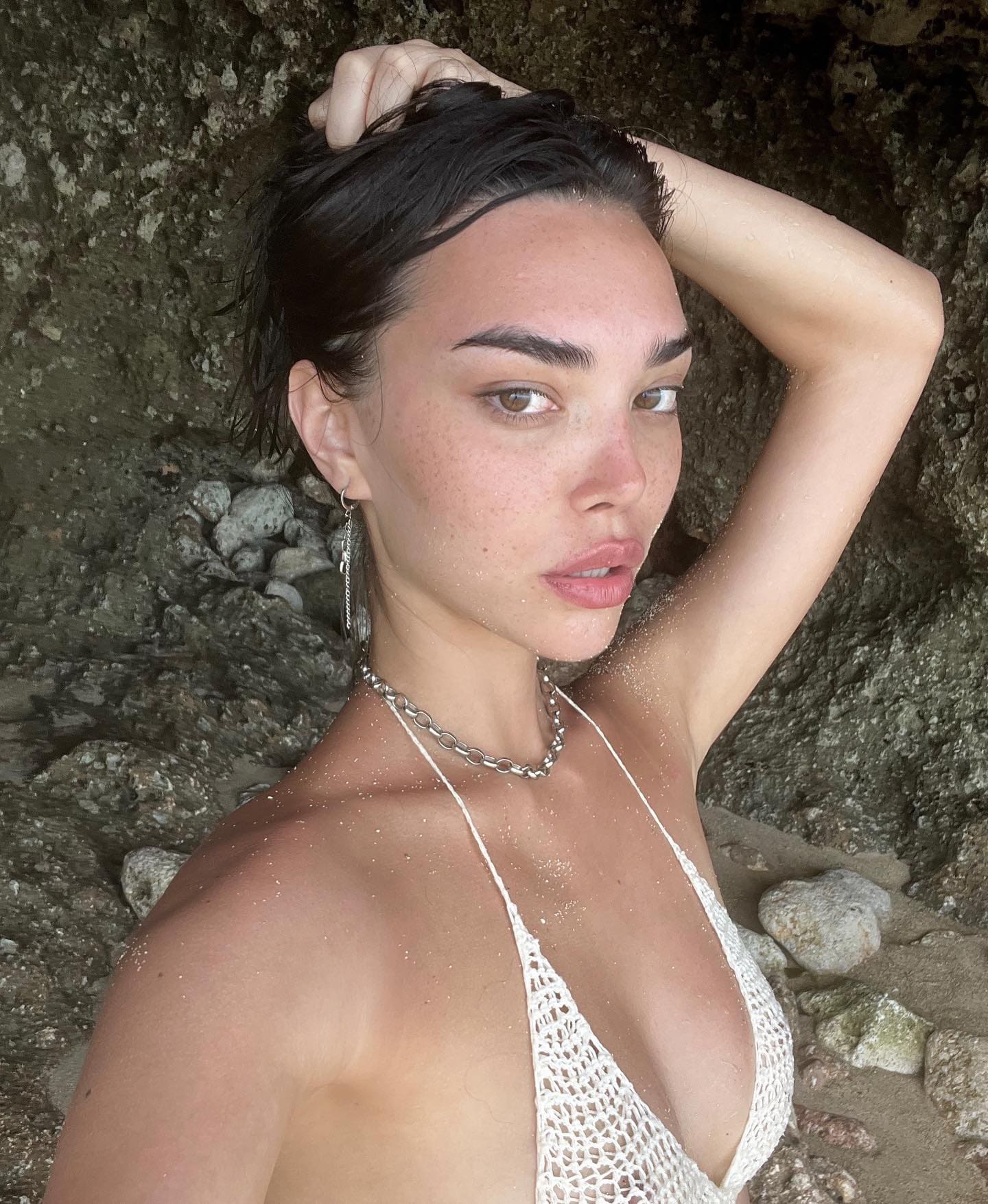 Adelina poses with hand on head, wearing a white crocheted halter top and silver necklace against a rocky background.