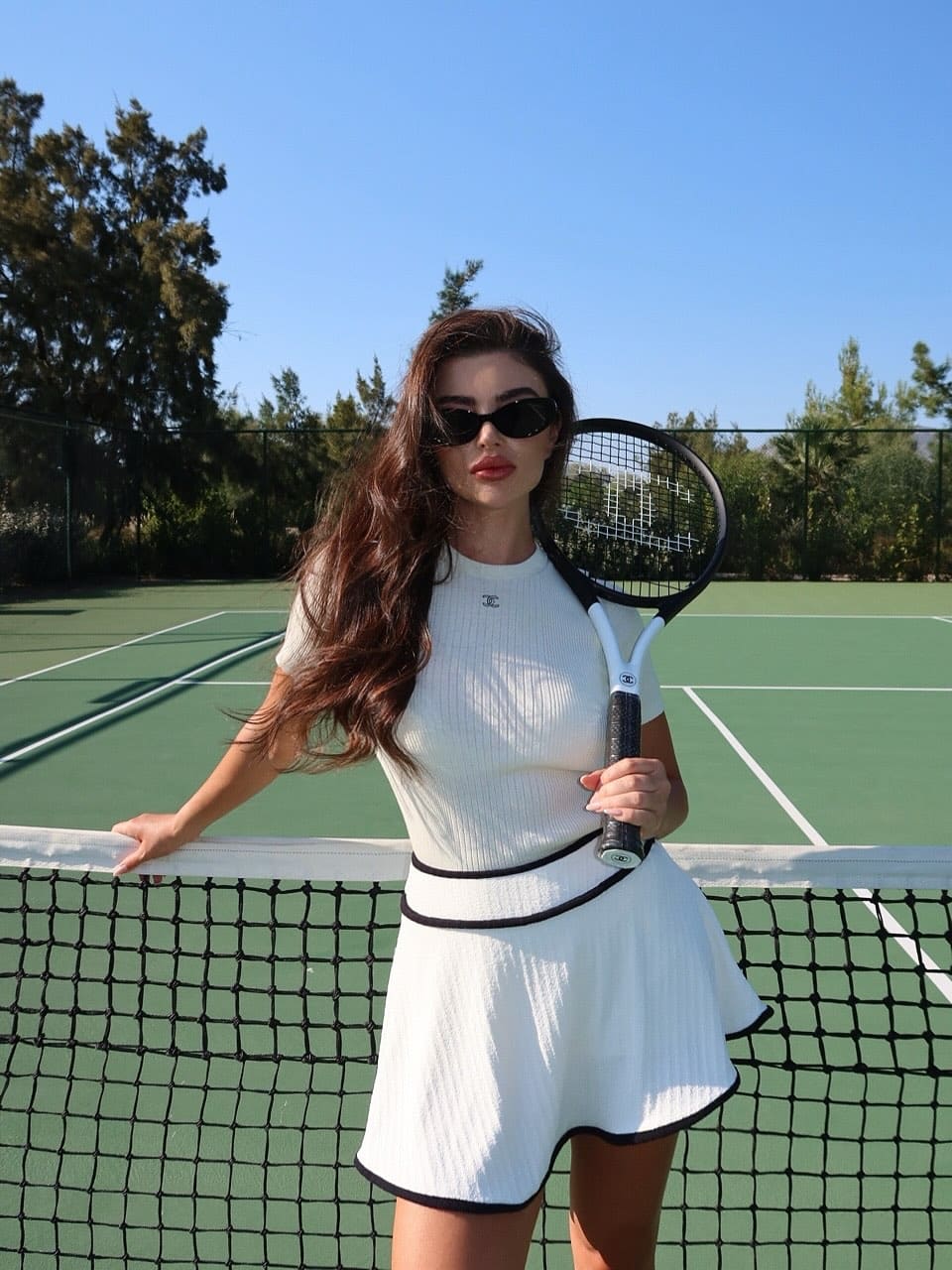 A woman stands on a tennis court in a white ribbed outfit and sunglasses, holding a racket upright and resting on the net.