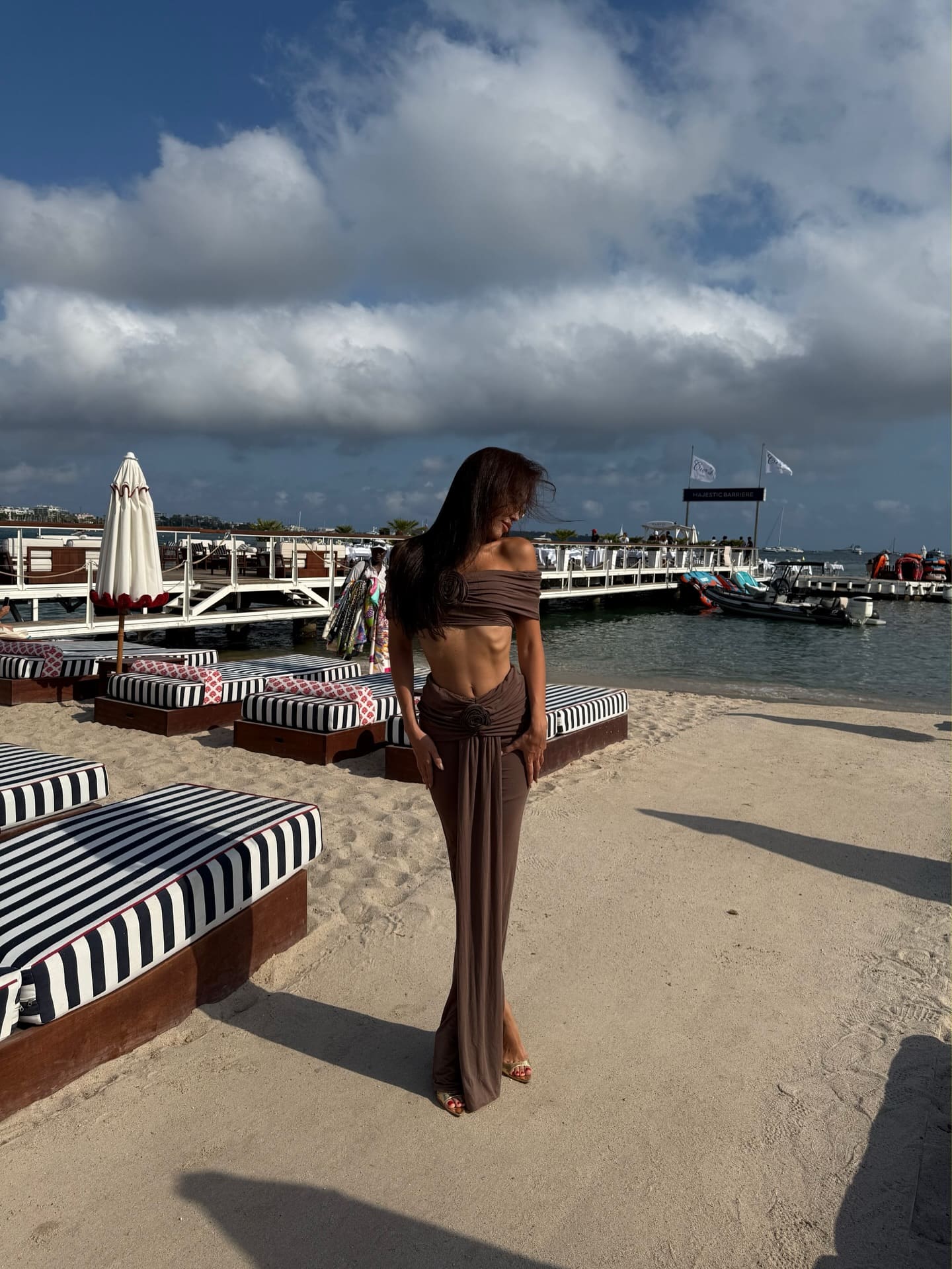 A woman stands on a sandy beach in a brown off-shoulder top and draped skirt, with striped lounge chairs and a pier in background.