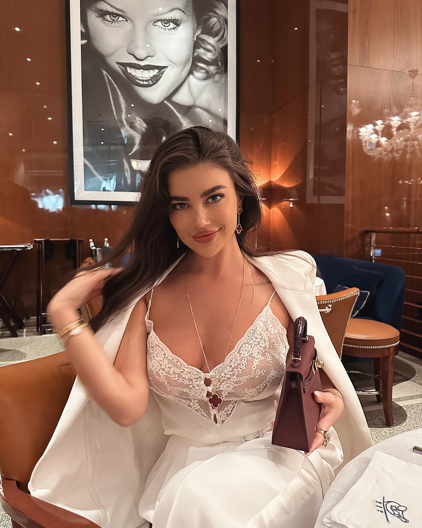 Aruzhan sits in a wood-paneled lounge wearing a white lace bustier and blazer, holding a maroon handbag.